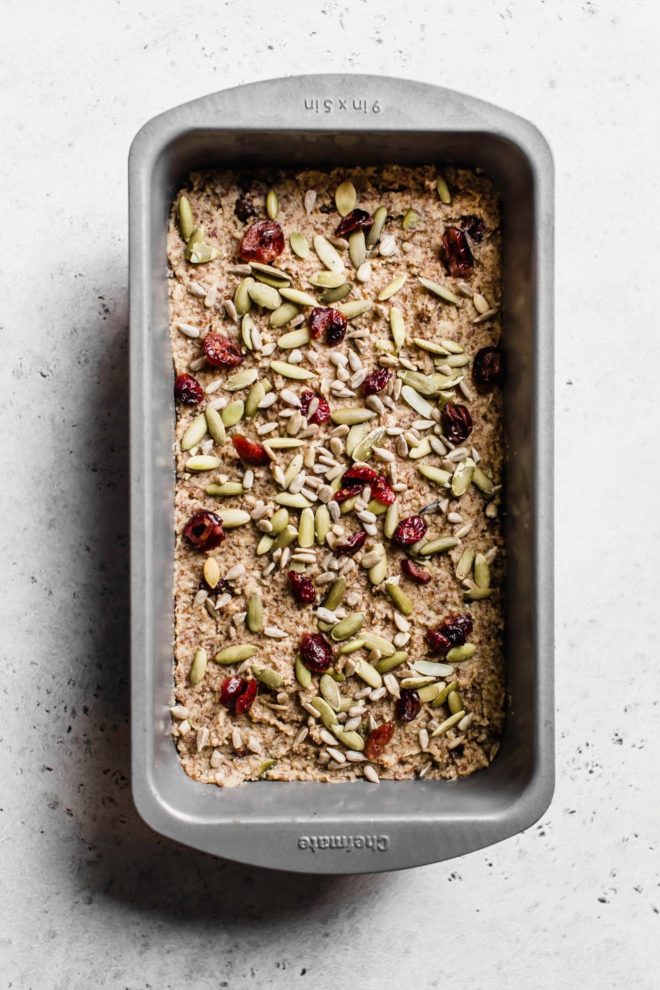 This is an overhead image of a bread pan with raw flaxseed bread batter in it. The batter is topped with sunflower seeds, pumpkin seeds, and dried cranberries. The loaf pan sits on a white surface.