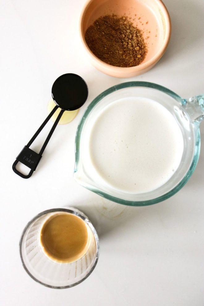 This is an overhead image of four items: a small pink bowl of spices, a tablespoon of agave nectar, a measuring cup of milk, and a small clear glass with espresso. The items sit on a white counter.
