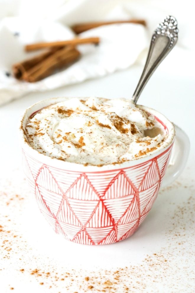 This is a side view of a white and red mug sitting on a white counter. The mug has a latte topped with whipped cream and a sprinkle of cinnamon. A white tea towel and cinnamon sticks are blurred in the background. An antique spoon is in the mug leaning against the side.