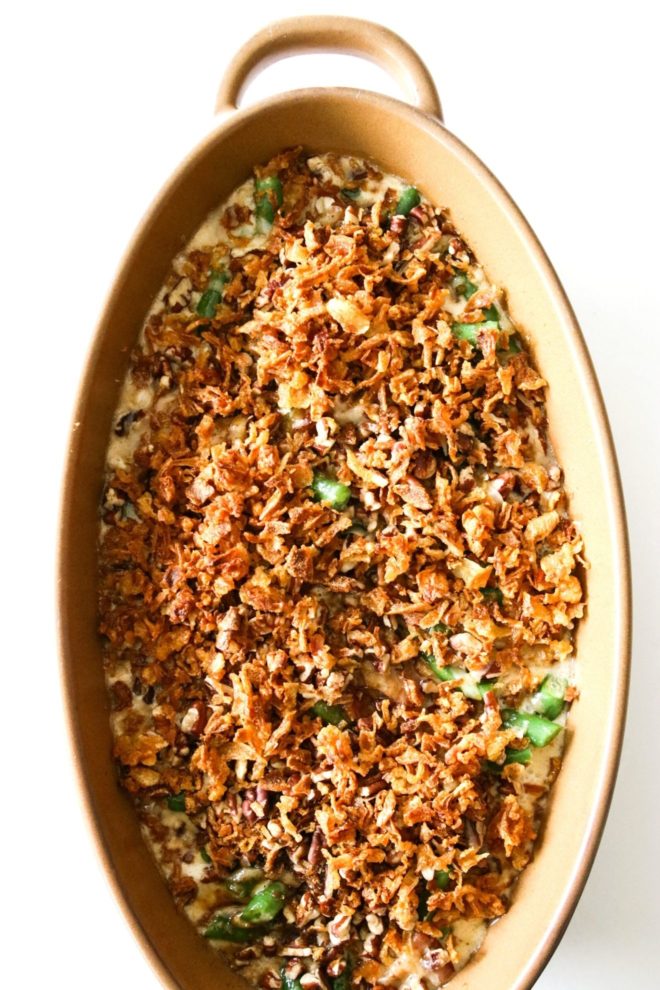 This is an overhead image looking into a light brown oval casserole dish with handles. The dish sits on a white counter. Inside the dish are green beans, cheese sauce, and fried crispy onions.