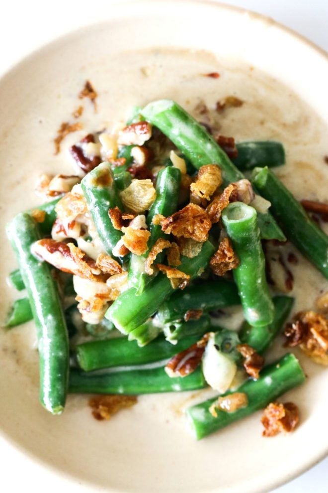 This is an overhead image looking down onto a tan plate with green beans smothered in a cheesy sauce and topped with fried onions and pecans.