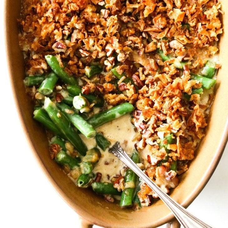 This is an overhead image looking down into an oval brown casserole dish with handles. The dish sits on a white counter. Inside the dish are cut green beans in a cheesy sauce topped with fried onions and pecans. An antique spoon is dipping into the green beans and leaning against the side of the dish.