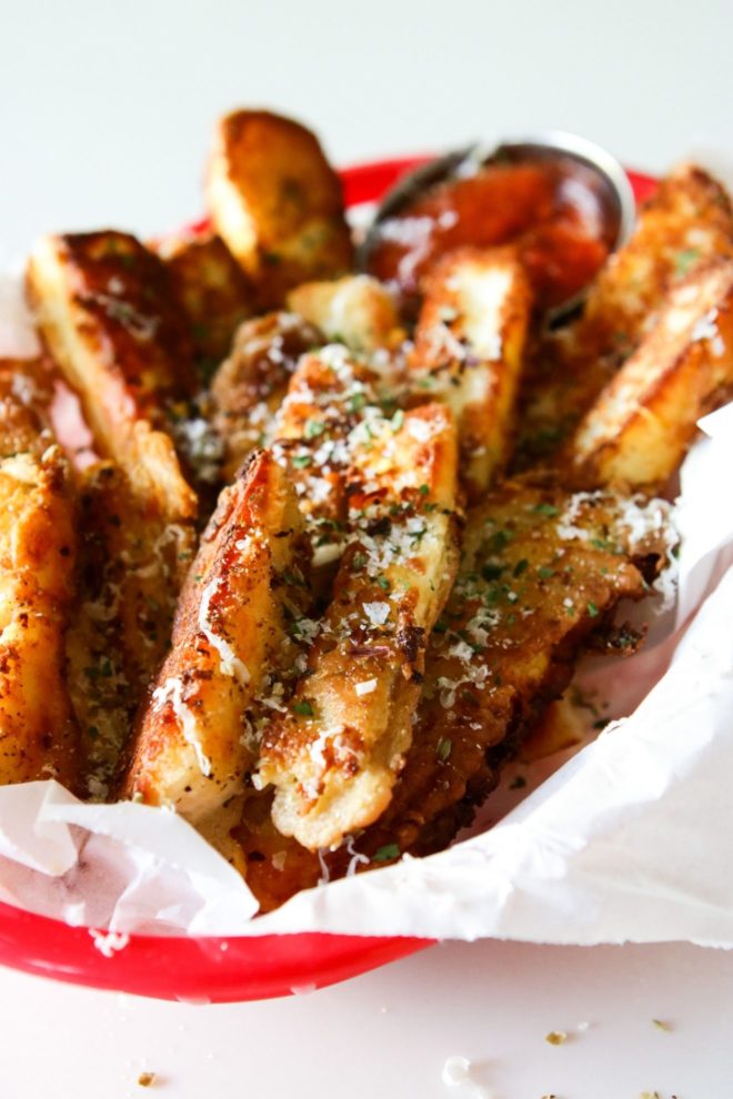 This is a side view closeup image of a red plastic basket lined with white parchment paper with halloumi fries in them. The fries are topped with grated cheese and dried oregano. A small silver metal bowl has marinara sauce in it and is blurred in the background. The basket sits on a white counter.
