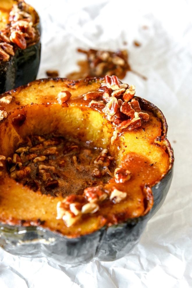 This is a side view of a cooked acorn squash cut in half and topped with a sweet pecan sauce. The acorn squash sits on a white piece of parchment paper.