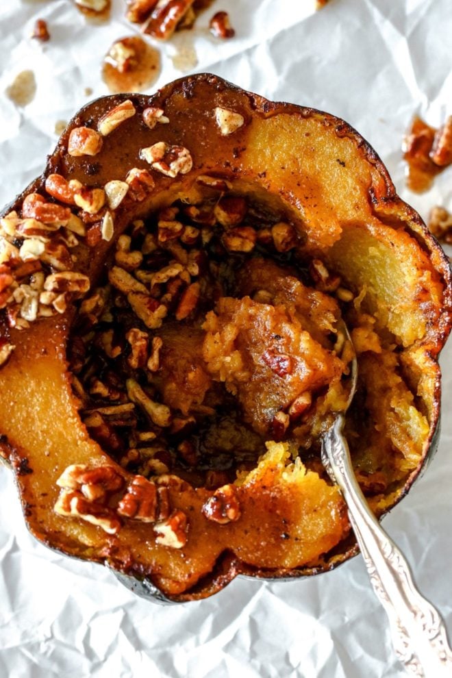 This is an overhead view of a cooked acorn squash with a spoon in it scooping up some orange flesh. The spoon is leaning against the side of the acorn squash. The squash is topped with pieces of pecans. The acorn squash sits on top of a piece of white parchment paper.