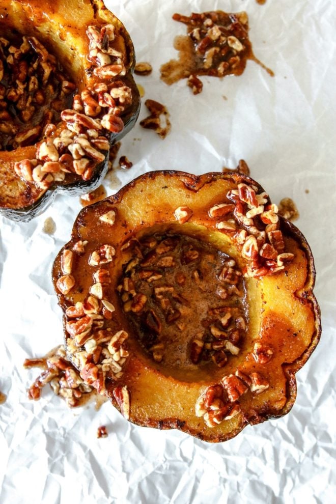 This is an overhead image of a cooked acorn squash cut in half. The acorn squash sits on a white piece of parchment paper and is topped with a cinnamon pecan sauce.