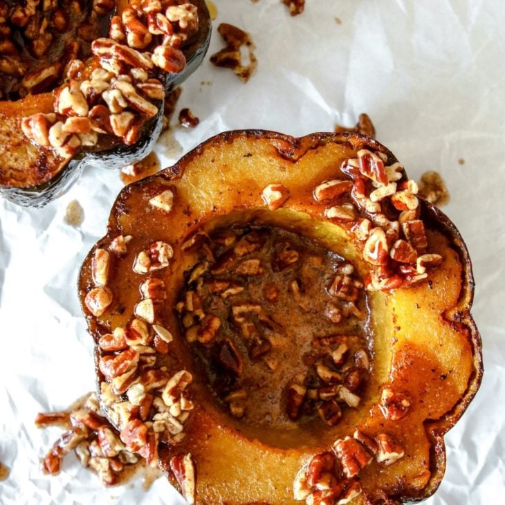 This is an overhead image of a cooked acorn squash cut in half. The acorn squash sits on a white piece of parchment paper and is topped with a cinnamon pecan sauce.