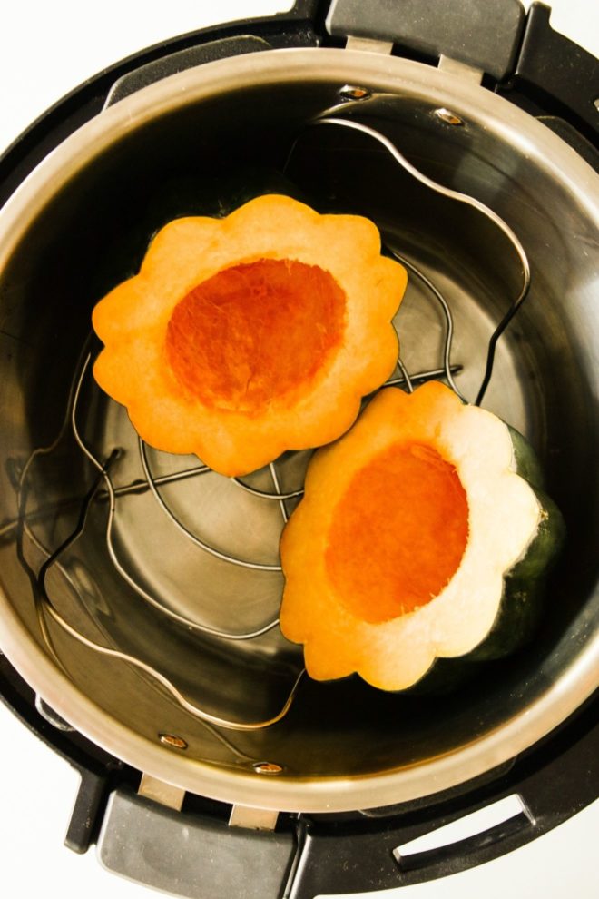 This is an overhead image looking into an instant pot with a metal trivet and an acorn squash cut in half. The instant pot sits on a white counter.