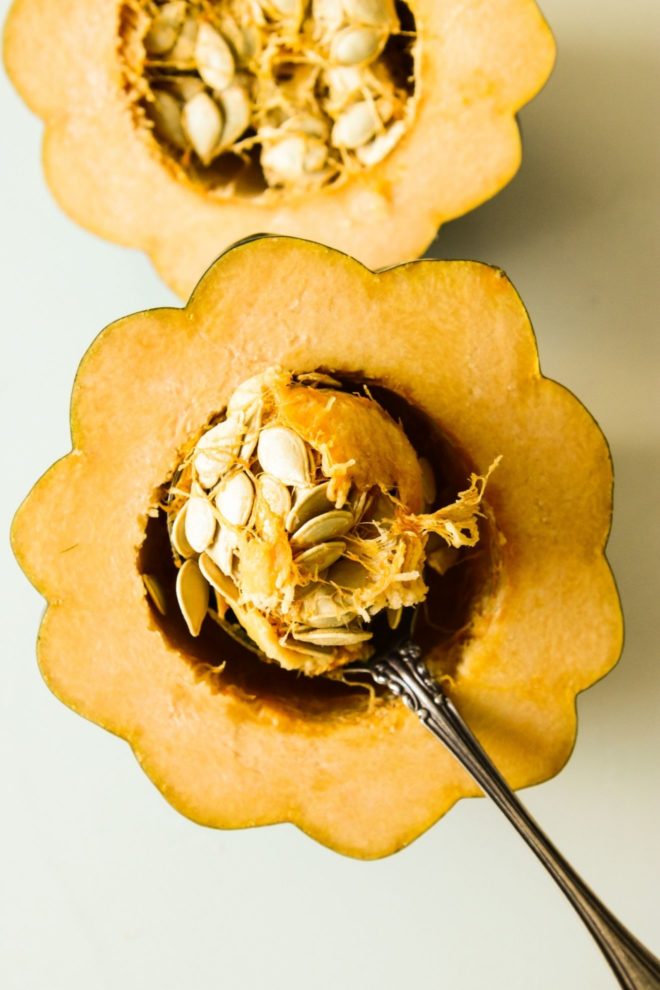 This is an overhead image of an acorn squash cut in half. An antique spoon is scooping the seeds out of the center. The acorn squash sits on a white counter.