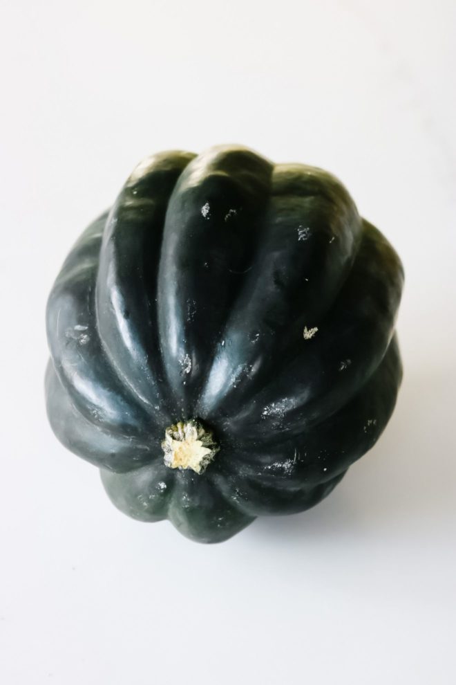 This is a side view of an acorn squash on a white counter. The stem is facing the camera and the ridges of the acorn squash are going backward.
