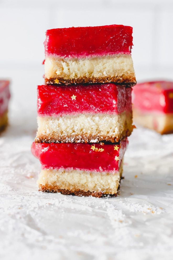 This is a stack of three cranberry lemon bars sitting on a white piece of parchment paper. More bars are blurred in the background. The bars have a shortbread base and bright magenta cranberry top layer.
