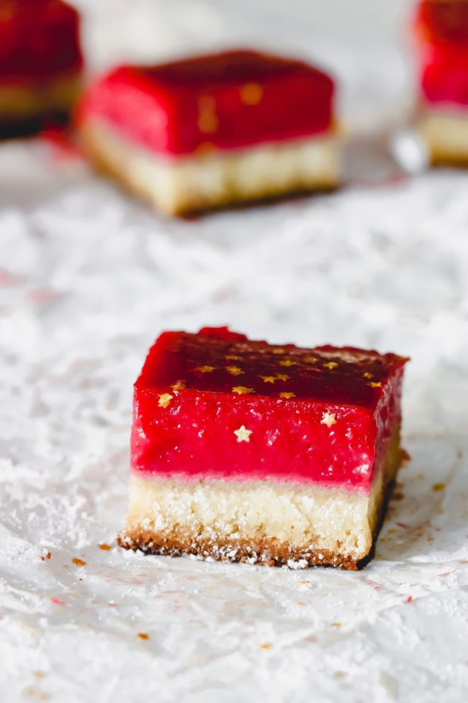This is a side view looking at a lemon cranberry bar on a white piece of parchment paper and more bars blurred in the background. The bar has a shortbread base and a bright magenta cranberry top layer. The bars are decorated with gold star sprinkles.