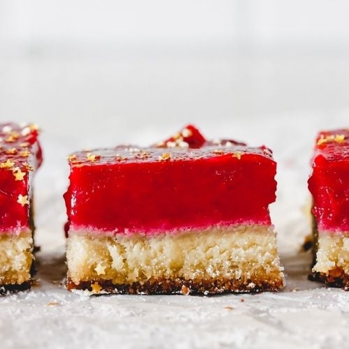 This is a side view of a lemon cranberry bar with more bars on either side of it. The bars sit on a white piece of parchment paper with a bite background. The cranberry lemon bars have a shortbread base and a bright magenta cranberry topping. The bars are topped with gold star sprinkles.