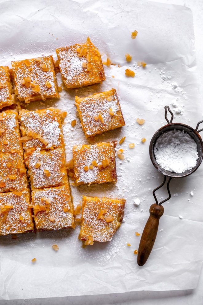 This is an overhead image of orange bars cut into squares on a white piece of parchment paper. The bars are topped with powdered sugar and candied oranges. An antique sifter is off to the right with powdered sugar in it.