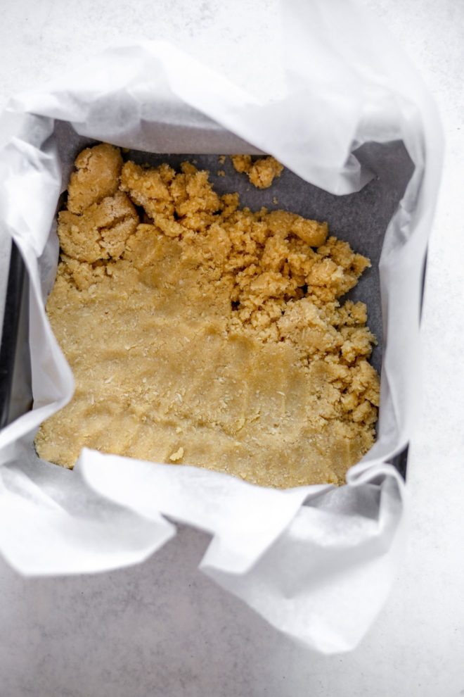 This is an overhead image of a square pan lined with parchment paper. The shortbread crust is halfway pressed into the bottom of the pan. The pan sits on a white surface.