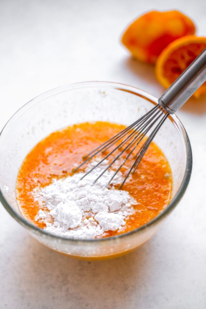This is a side view looking into a glass bowl with an orange juice mixture with arrowroot flour. The bowl sits on a white surface with squeezed orange halves blurred in the background.