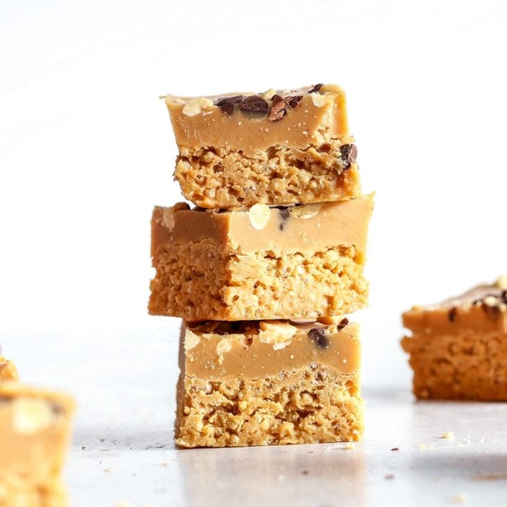 This is a stack of three small peanut butter squares. Each square has a top creamy layer and a bottom crunchy layer. The stack sits on a white surface with more squares blurred in the background.