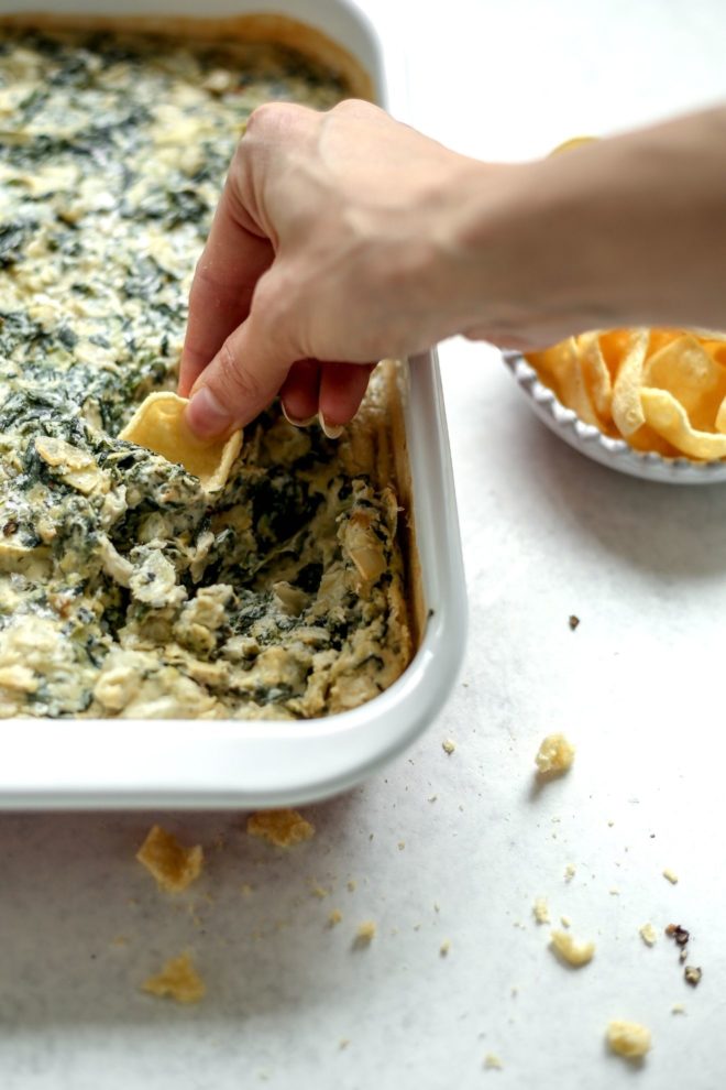 This image is peering into a white baking dish with spinach artichoke dip. A hand is holding a chip on the right side of the image and scooping up some of the dip. The baking dish sits on a white surface with a small bowl of chips to the side of it.