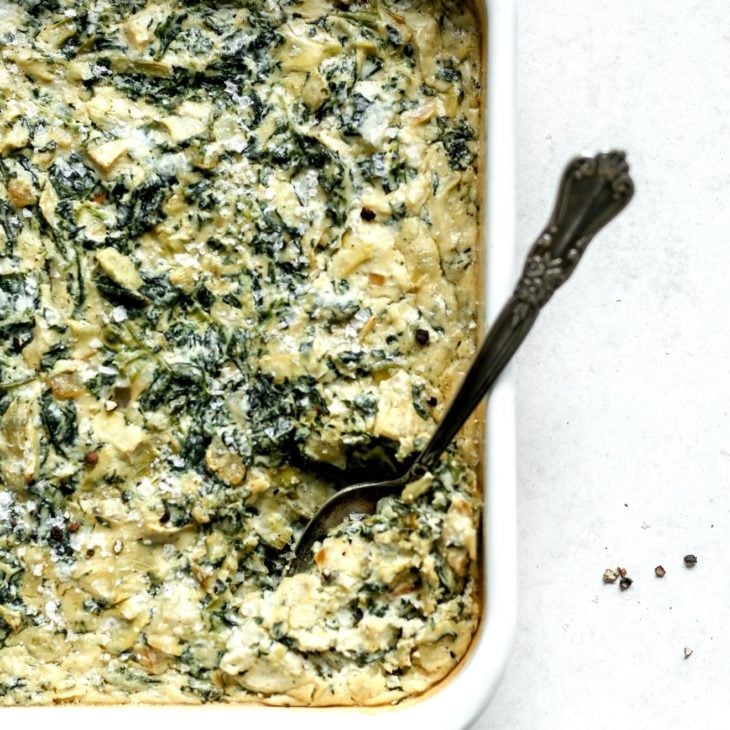 This is an overhead image of a white baking dish with vegan spinach artichoke dip. A spoon is scooping up some dip and leaning on the side of the dish. The dish sits on a white surface.