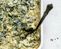 This is an overhead image of a white baking dish with vegan spinach artichoke dip. A spoon is scooping up some dip and leaning on the side of the dish. The dish sits on a white surface. Text overlay reads "vegan spinach artichoke dip get the recipe at thetoastedpinenut.com"