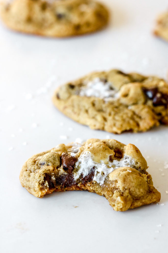 This is a side view of a sourdough cookies with chocolate chips and marshmallows. The cookie has a bite taken out of it and sits on a white counter. More cookies are blurred in the background.
