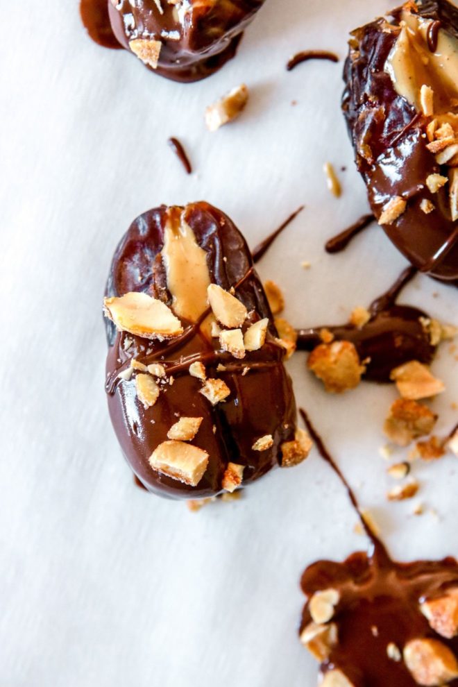 This is a close up of dates covered in chocolate and stuffed with peanut butter. The dates are sprinkled with chopped peanuts. The dates sit on a white surface.