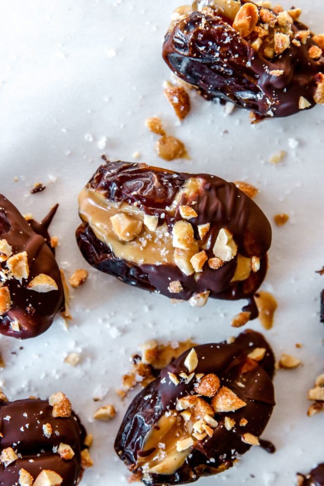 This is an overhead image of chocolate coated dates stuffed with peanut butter and sprinkled with chopped peanuts and salt. The dates sit on a white piece of parchment paper.