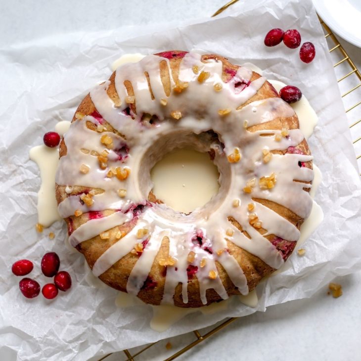 This is an overhead image of a cranberry orange cake glazed with a white icing and candied orange pieces. The cake sits on a white piece of parchment paper on a cooling rack. The cooling rack sits on a white surface. A few fresh cranberries are sitting on the parchment paper next to the cake. A small white plate with silver utensils is next to the cooling rack to the top right corner of the image.