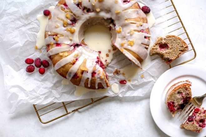 This is an overhead image of a cranberry orange cake glazed with a white icing and candied orange pieces. The cake sits on a white piece of parchment paper on a cooling rack. The cooling rack sits on a white surface. A few fresh cranberries are sitting on the parchment paper next to the cake. A slice of cake is on the cooling rack next to the cake and on a white plate to the bottom right of the image.
