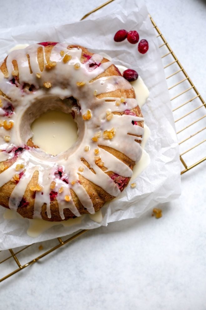 This is an overhead image of a cranberry orange cake glazed with a white icing and candied orange pieces. The cake sits on a white piece of parchment paper on a cooling rack. The cooling rack sits on a white surface. A few fresh cranberries are sitting on the parchment paper next to the cake.