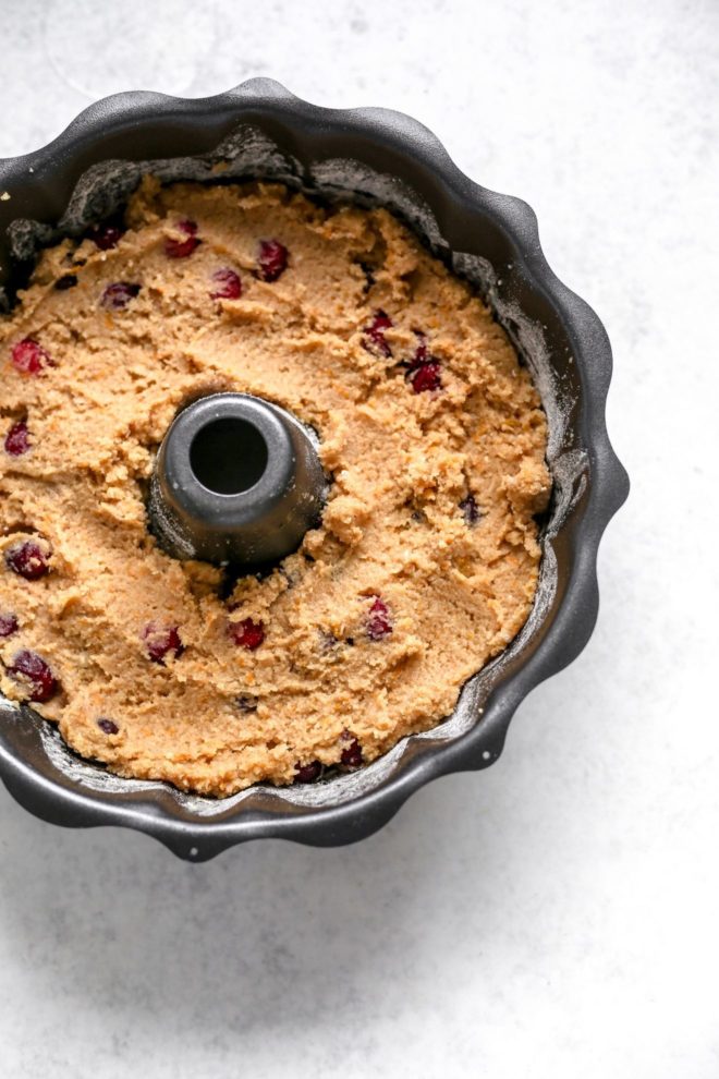 This is an overhead image of a bundt pan with cake batter in it. The pan sits on a white surface. The batter is a tan/beige color with fresh cranberries mixed in.