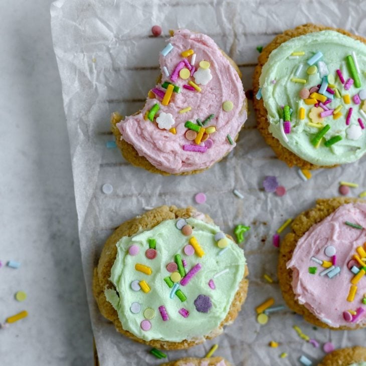 This is an overhead image of a cooling rack coming from the lefthand side of the image. The cooling rack sits on a light grey surface with a white piece of parchment paper on top. On the parchment paper is six lofthouse cookies with pink and light green icing and rainbow sprinkles. The cookie to the top left has a bite taken out of it.