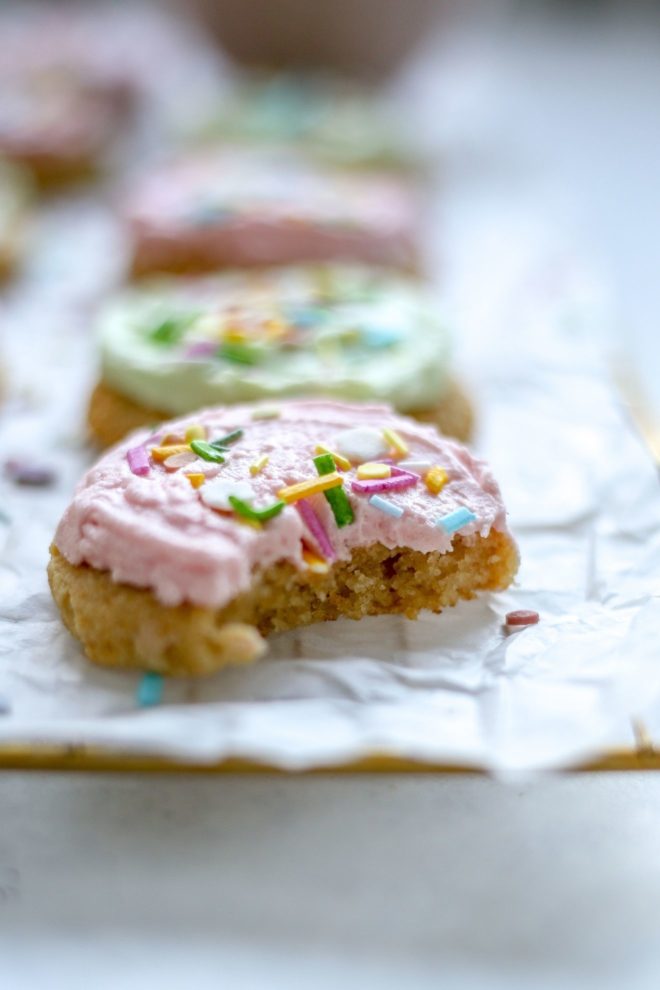 This is a side view of a lofthouse cookie with pink frosting and rainbow sprinkles with a bite taken out of it. The cookie sits on top of a white piece of parchment paper on a cooling rack on a white counter. More lofthouse cookies with light green and pink frosting are blurred in the background.