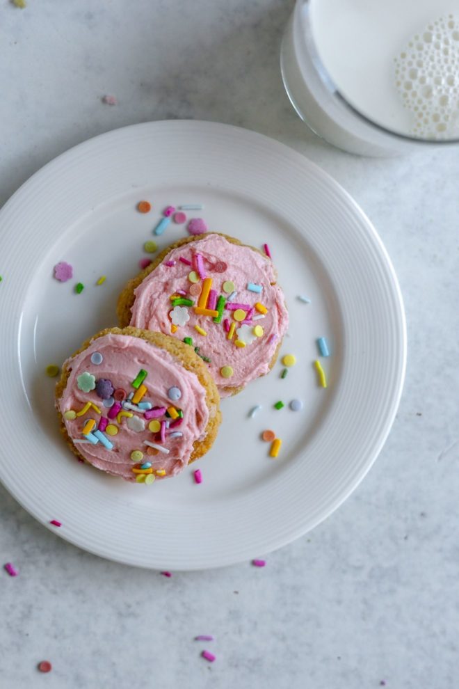 This is an overhead image of a small white plate with two lofthouse cookies with pink frosting. The cookies are topped with rainbow sprinkles The plate sits on a light grey surface and a glass of milk is to the top right of the image.