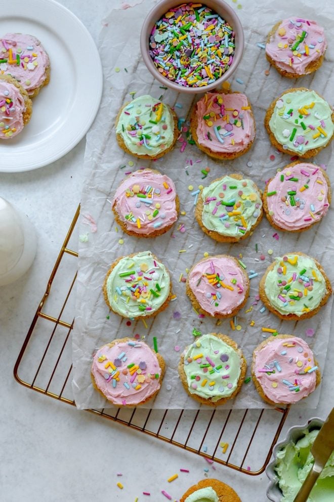 This is an overhead image of a cooling rack with a piece of white parchment paper on it. On the parchment paper is 13 lofthouse cookies with pink and green frosting and colorful sprinkles. To the bottom of the image is a small bowl with light green frosting and to the left top of the image is a small white plate with two cookies with pink frosting and a glass of milk.