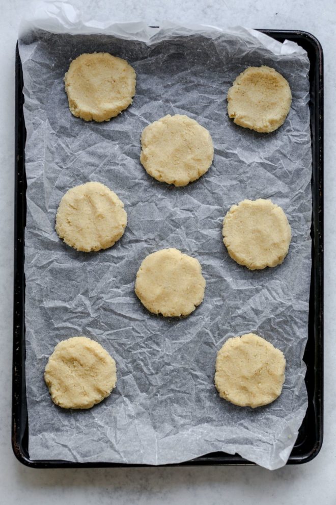 This is an overhead image of a bakinf sheet with parchment paper and 8 raw cookie dough circles on top. The baking sheet sits on a light grey surface.
