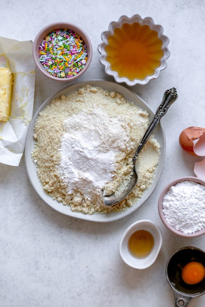 This is an overhead image of butter, sprinkles, honey, almond flour, coconut sugar, eggs, powder sugar and vanilla in separate bowls on a white counter.