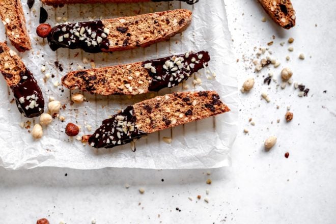 This is an overhead view of biscotti dipped in chocolate and sprinkled with chopped hazelnuts. The biscotti lays on a white piece of parchment paper on a cooling rack. The cooling rack sits on a white surface. One biscotti is to the right side of a cooling rack on the white surface. Some chopped hazelnuts are on the surface next to the biscotti.