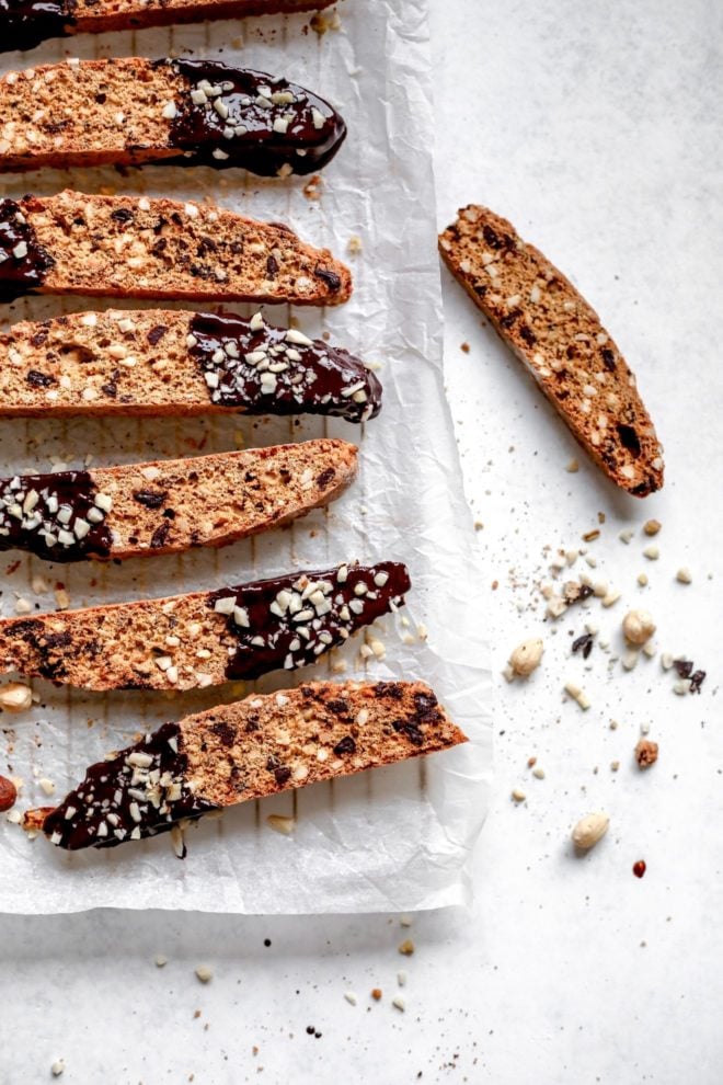 This is an overhead view of biscotti dipped in chocolate and sprinkled with chopped hazelnuts. The biscotti lays on a white piece of parchment paper on a cooling rack. The cooling rack sits on a white surface. One biscotti is to the right side of a cooling rack on the white surface. Some chopped hazelnuts are on the surface next to the biscotti.