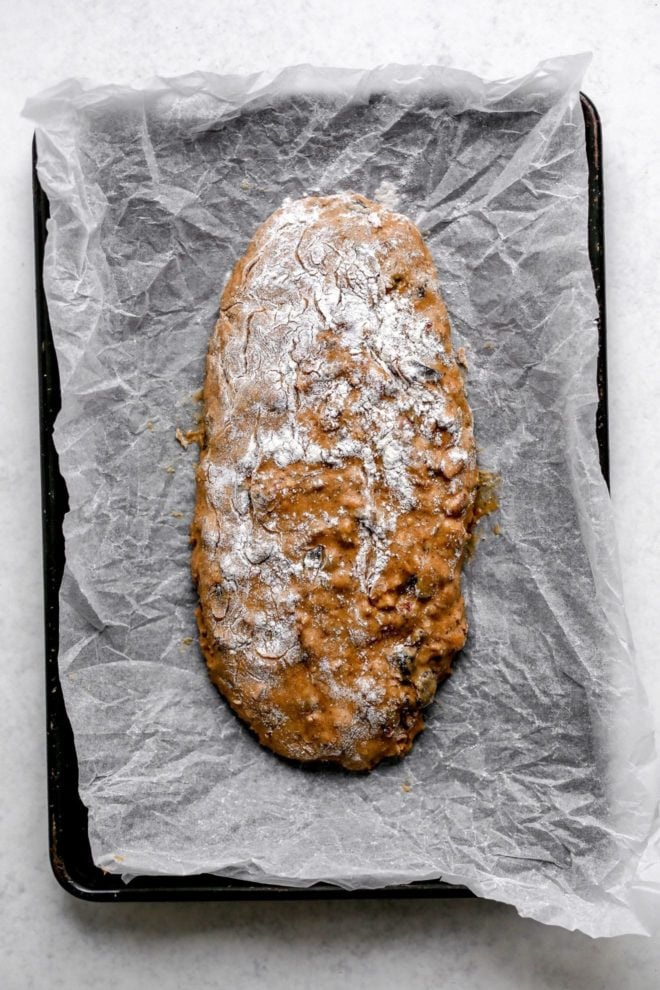 This is an overhead image of biscotti dough shaped in a large rectangle log on a piece of parchment paper on a baking sheet. The baking sheet sits on a white surface.