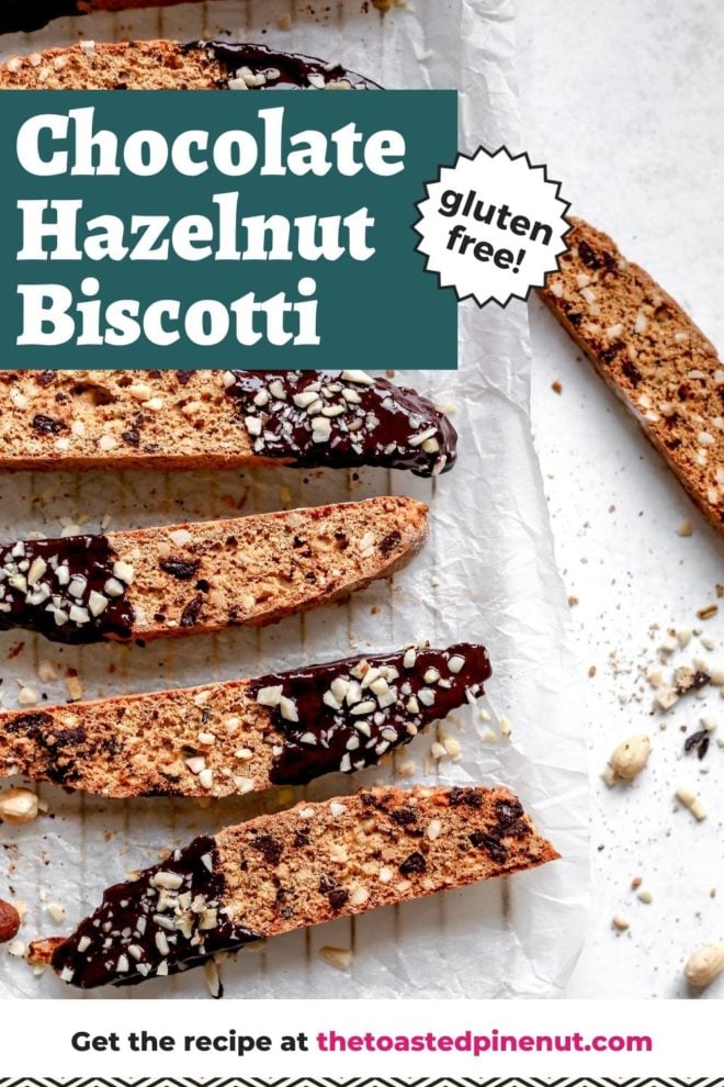 This is an overhead view of biscotti dipped in chocolate and sprinkled with chopped hazelnuts. The biscotti lays on a white piece of parchment paper on a cooling rack. The cooling rack sits on a white surface. One biscotti is to the right side of a cooling rack on the white surface. Some chopped hazelnuts are on the surface next to the biscotti. Text overlay reads "chocolate hazelnut biscotti gluten free!"