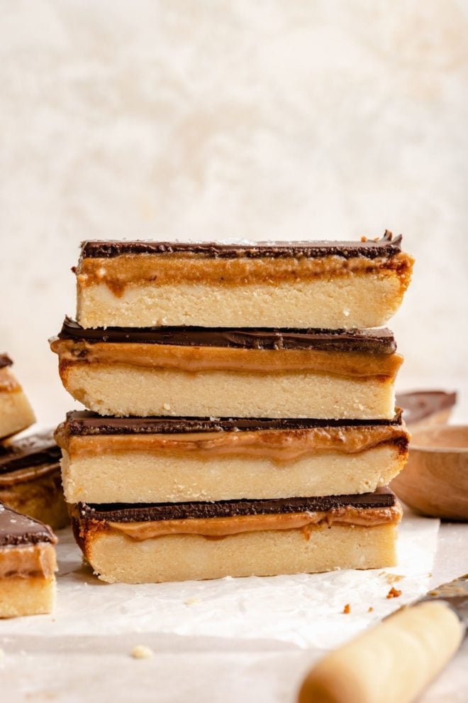 This is a stack of four twix bars. You can see the layers of the twix bars. The stacks sits on a white piece of parchment paper on a beige surface with more twix bars around the stack.