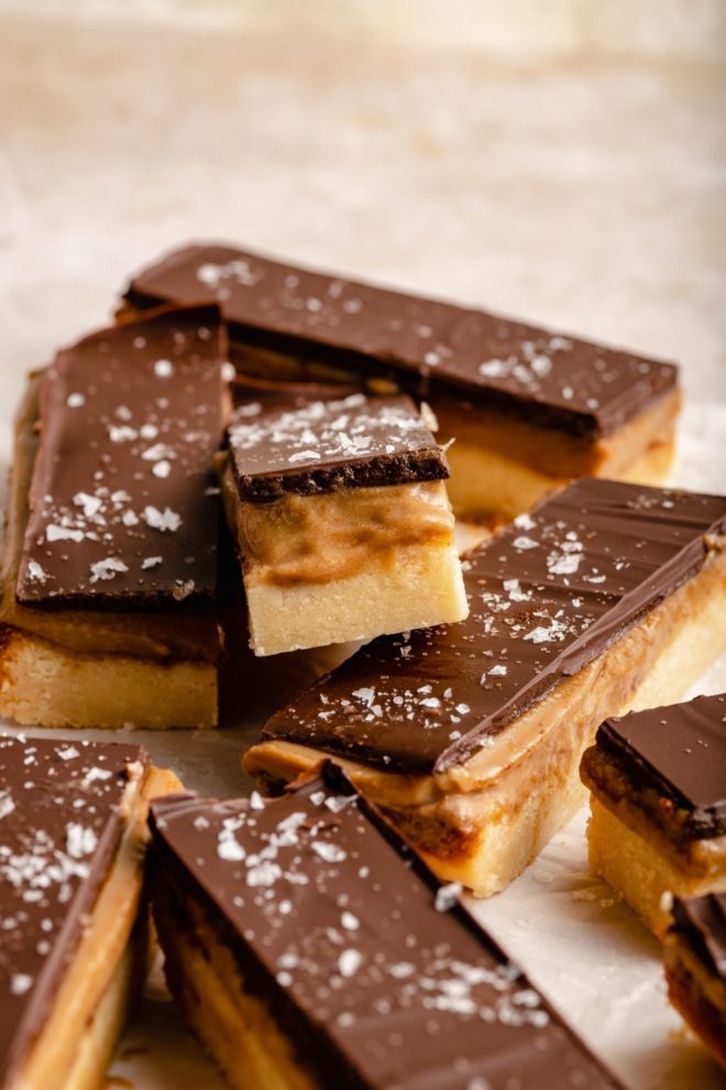 This is a closeup of a twix bar with three layers: shortbread, caramel, and chocolate. The bar is leaning against the other bars. The bars sit on a white/beige surface.