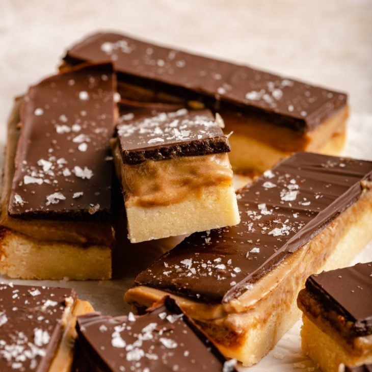This is a closeup of a twix bar with three layers: shortbread, caramel, and chocolate. The bar is leaning against the other bars. The bars sit on a white/beige surface.
