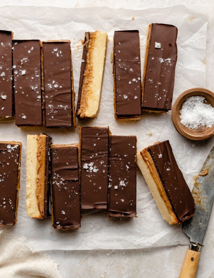 This is an overhead image of homemade twix bars lined up on a white piece of parchment paper. A couple twix bars are turned to the side to reveal the layers. A small bowl of salt is to the right side of the image, next to the bars. A knife is laying on its side next to the salt bowl and twix bars.