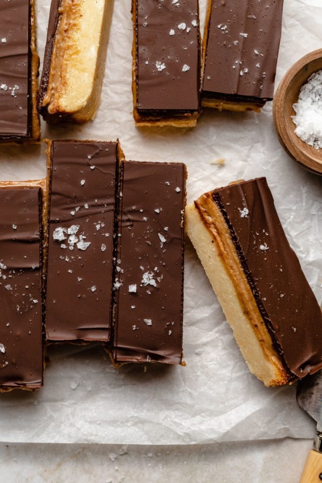 This is an overhead image of homemade twix bars lined up on a white piece of parchment paper. A couple twix bars are turned to the side to reveal the layers.