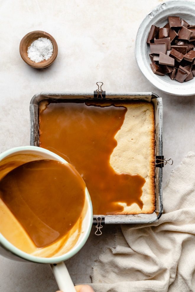 This is an overhead image of a square pan with a cooked shortbread layer. A pan with caramel sauce is being pouring on top of the shortbread layer. The pan sits on a white surface and a small bowl of chocolate is to the top right corner of the image. A beige tea towel is to the bottom corner. A small bowl of salt is to the top left corner of the image.