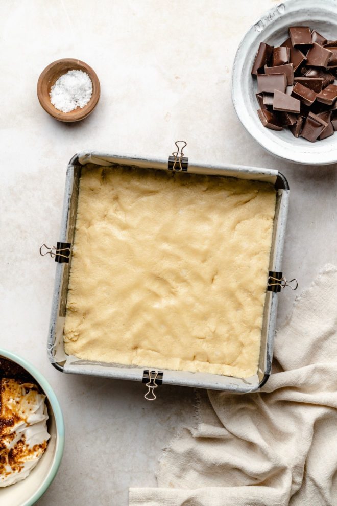 This is an overhead image of a square pan with uncooked shortbread layer at the bottom. The square pan sits on a light grey surface with a beige tea towel to the bottom right corner, a small bowl of chocolate to the top right of the image, a small bowl of salt to the top left corner.