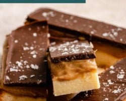 This is a closeup of a twix bar with three layers: shortbread, caramel, and chocolate. The bar is leaning against the other bars. The bars sit on a white/beige surface. Text overlay reads "homemade twix bars vegan & gluten free!"
