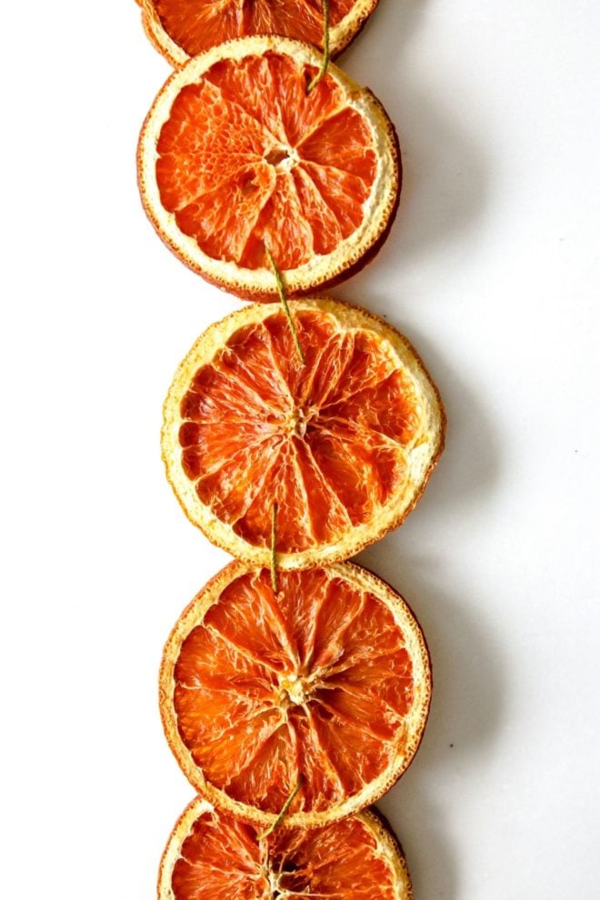 This is a closeup of dried orange slices strung together with a green string. The orange slices are vertical in the middle of the image with the white surface on either side.
